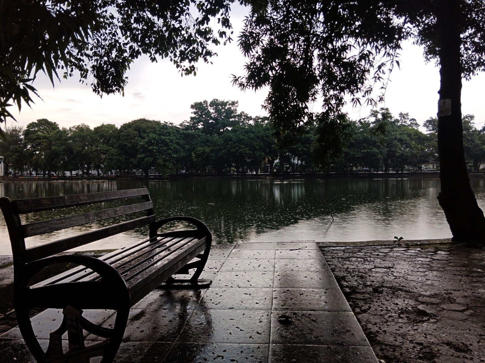 Danau Taman Kambang Iwak Palembang dengan bangku taman di tepi air saat hujan